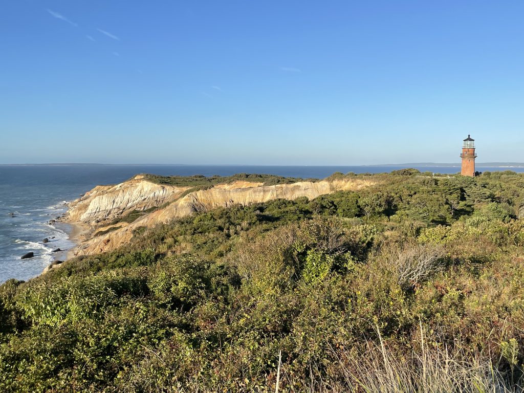 A view of long, craggy, sandy cliffs in the distance and a red brick lighthouse nearby.